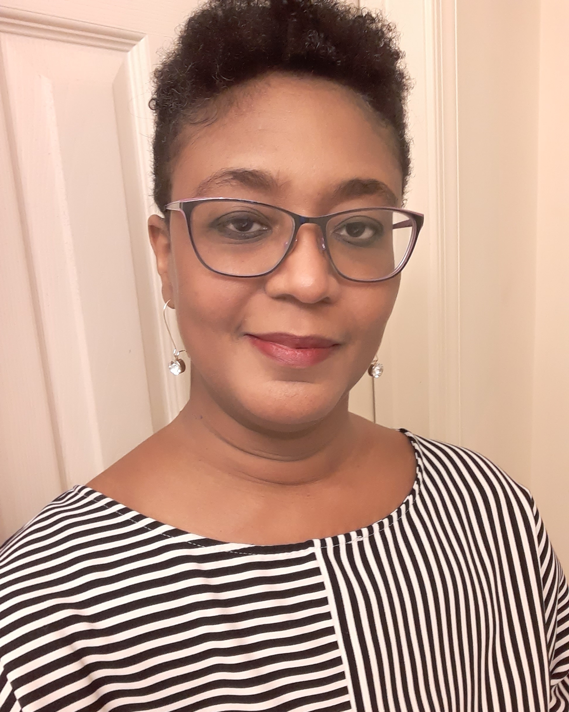 
		A close-up of Camisha Jones smiling slightly. She is a Black cis-woman with gingerbread brown skin. Her dark brown naturally textured hair is short, peppered with silver strands, and cut close on the sides in a fade style. She wears black and purple-framed glasses, ruby red lipstick, dangling earrings with a clear colored jewel at the end, and a black and white top with horizontal stripes on one side and vertical stripes on the other.
		