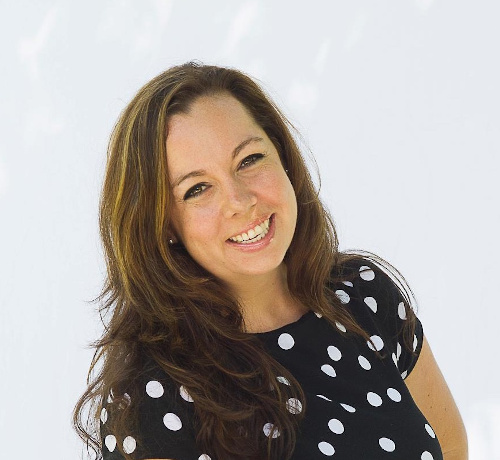 
		Mariclare wears a black and white polka dotted dress in a brightly lit room. A white woman with wavy brown hair, brown eyes, and glossed pink lips, she smiles and tilts her head slightly off to her right. 
		