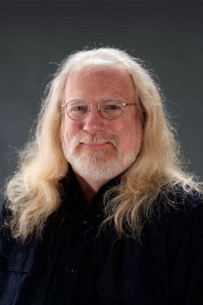 
	Michael Maag headshot. He is a white man with flowing white/blonde hair and a short beard. He wears wire rimmed glasses and a dark blue collared shirt as he smiles warmly. Photo Robbie Sweeny.
		