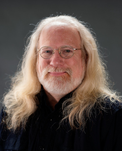 
	Michael Maag headshot. He is a white man with flowing white/blonde hair and a short beard. He wears wire rimmed glasses and a dark blue collared shirt as he smiles warmly. Photo Robbie Sweeny.
		