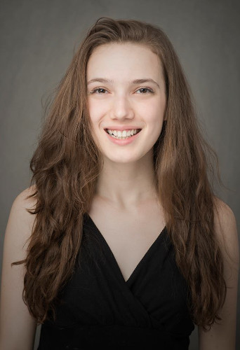 
		A portrait photo of a white woman in a black V-neck tank top with a grey background. Her long semi-wavy light brown hair falls symmetrically next to each arm as her body and big smile aim directly to the camera.
		