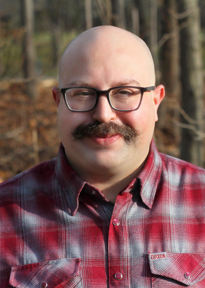 
	Matt is a white man with round face, shaved head, bushy brown mustache, and dark-rimmed glasses.  Clad in a plaid shirt and backed by trees, his eyes crinkle as he smiles cheerfully and mischieviously at you.	
	