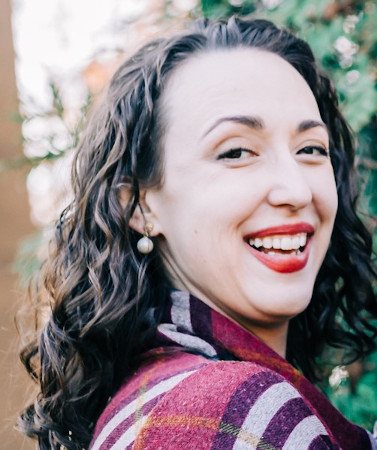 Stephanie looks back over her right shoulder, smiling widely. She is a white woman with wavy brown hair and sharp eyebrows; she's sporting bright red lipstick and a purple striped jacket. 